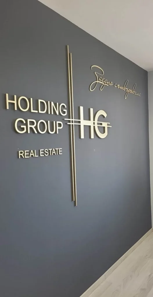 Обемно златисто лого HOLDING GROUP HG с ръкописен подпис на сива стена в офис